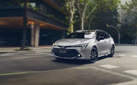 Srebrna Toyota Corolla kombi z czarnymi felgami sunie po miejskiej ulicy, otoczona zielenią i nowoczesną architekturą.