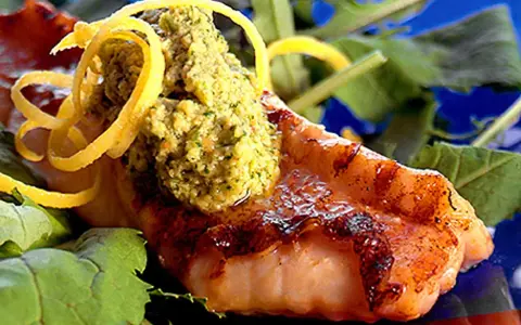 Grillad havsabborre med en grön topping och citronskal, serverad på en bädd av gröna blad.