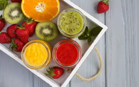 Bandeja con fresas, rodajas de kiwi, naranja y tres tarros de **puré de frutas** de colores vibrantes: verde, amarillo y rojo.