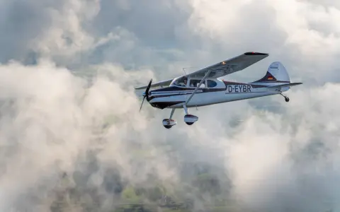 Eine braun-weiße Cessna 170 fliegt durch die Wolken über grünen Feldern.