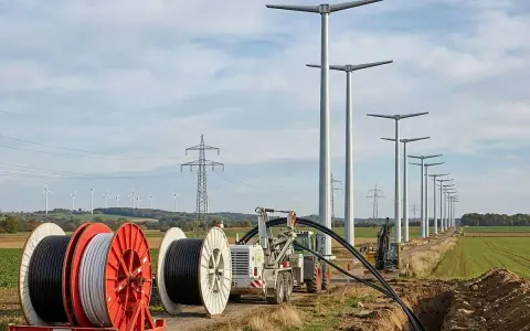 Kabeltrommeln und Bagger im Feld, während neue Windräder aufgestellt werden. Die Energiewende Herausforderungen bei der Infrastruktur sind sichtbar.