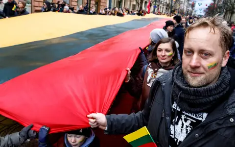 Ludzie niosą ogromną flagę Litwy podczas święta narodowego. Na twarzach uczestników widać patriotyczne barwy.