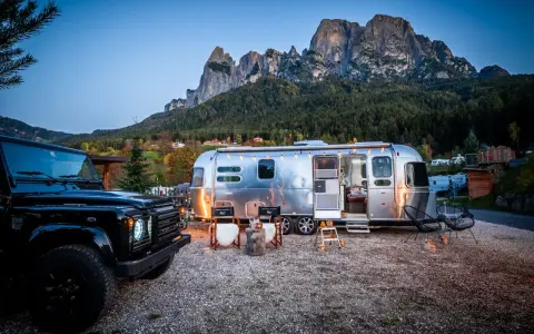 Ein Airstream-Wohnwagen und ein schwarzer Geländewagen stehen auf einem **Campingplatz Dolomiten** mit majestätischen Bergen im Hintergrund. Gemütliche Stühle und Lichter schaffen eine einladende Atmosphäre.