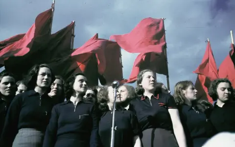 En grupp kvinnor deltar i ett 1 maj tåg, med röda fanor vajande i bakgrunden.