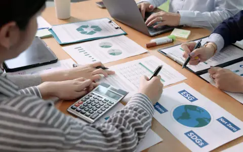 Des mains travaillent sur des documents financiers, une calculatrice et un ordinateur portable. L'ergonomie organisationnelle est au cœur de cette réunion axée sur le développement durable.