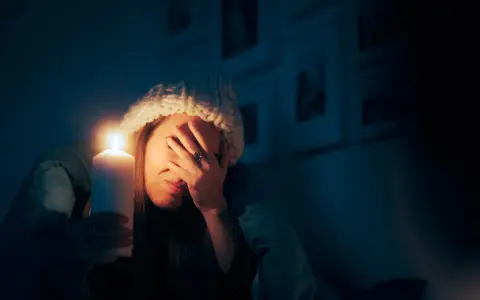 Kobieta w czapce trzyma świecę, zasłaniając twarz. To skutki przerwania dostaw energii elektrycznej do urządzeń o kluczowym znaczeniu.