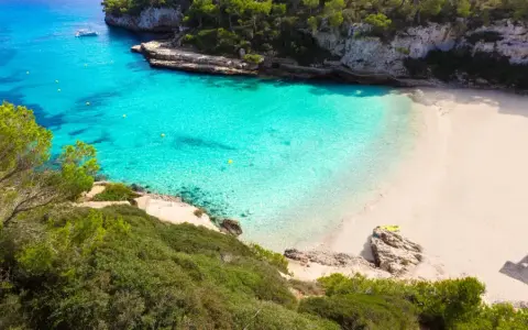 Traumhafte mallorca buchten mit türkisfarbenem Wasser, weißem Sand und grünen Pinien. Ein Boot ankert im Meer, eine Rettungsschwimmerturmbefindet sich am Strand.