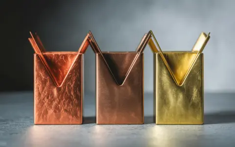 Trois porte-stylos aux formes géométriques, aux teintes cuivrées et laiton couleur, sur un fond gris.