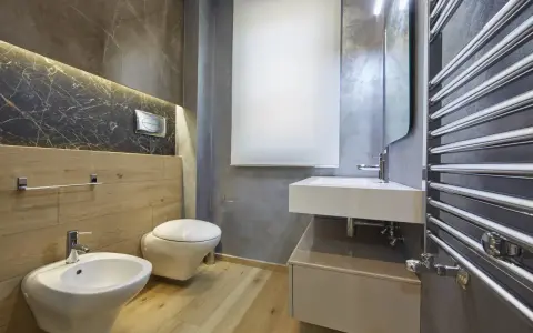 Bagno moderno con bidet, WC sospeso, lavabo e scaldasalviette cromato. L'illuminazione a LED sotto il marmo crea un'atmosfera elegante, rispettando l'altezza minima soffitto.