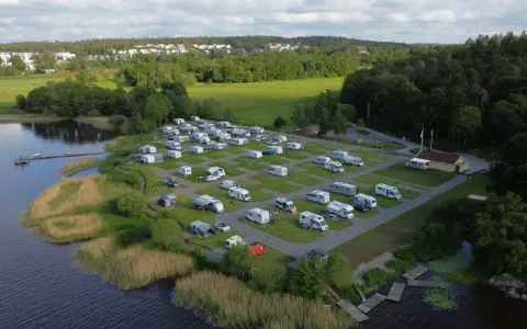 Full campingplats med husbilar och husvagnar vid en sjö, med grönskande träd och bebyggelse i bakgrunden. En idyllisk plats för camping i Alingsås.