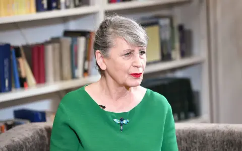 Joanna Wnuk-Nazarowa w zielonej bluzce z broszką w kształcie ważki, na tle regałów z książkami.