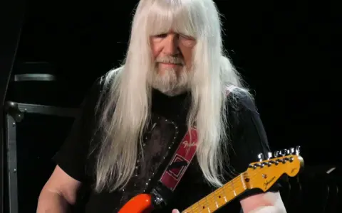 Andy Scott mit seinen charakteristischen langen, weißen Haaren spielt auf einer orangen E-Gitarre.