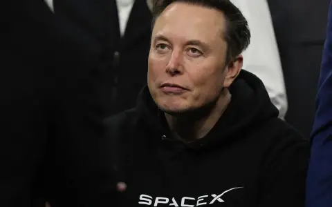 Elon Musk w bluzie SpaceX, zamyślony, w otoczeniu innych osób.