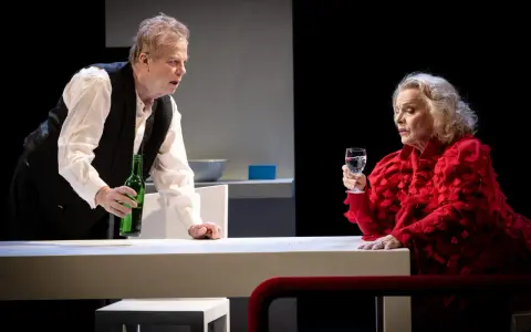 Krister Henriksson i en dramatisk scen. Han håller en grön flaska, medan en kvinna i rött med ett glas vatten ser på. Scenen påminner om hans roller i filmer och tv-program.