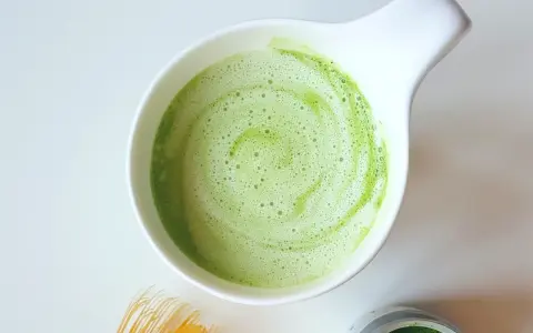 Zielony, spieniony napój matcha w białej miseczce, obok bambusowa miotełka i puszka z proszkiem. Idealne co na pobudzenie zamiast kawy.