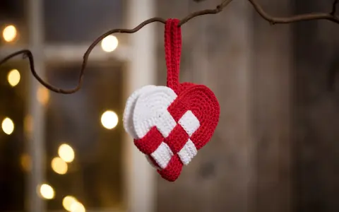 Ett virkat julhjärta i rött och vitt hänger från en gren, med julbelysning i bakgrunden.