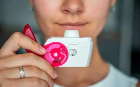Nahaufnahme einer Person, die einen Pulverinhalator hält, bereit zur Anwendung. Der Inhalator ist weiß mit pinken Akzenten und zeigt die Dosis "27".