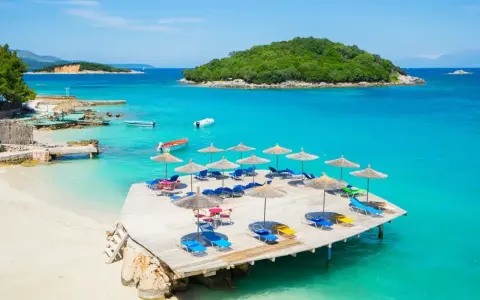 Rajskie ksamil plaże z leżakami i parasolami, turkusowa woda, zielona wyspa w tle.