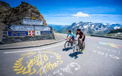 Ciclisti in salita verso il Col du Galibier, con montagne innevate sullo sfondo e segnaletica stradale.