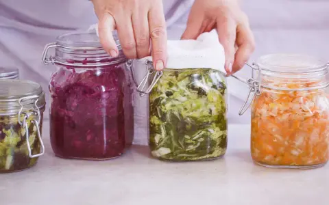 Hände bedecken ein Glas mit fermentiertem Gemüse, Teil einer Auswahl an probiotischen Lebensmitteln in Gläsern.