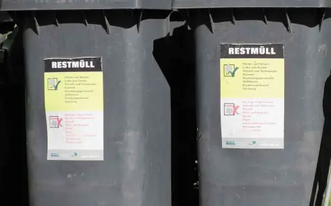 Dwa szare kosze na śmieci z etykietami "REST MÜLL". Etykiety zawierają informacje o tym, co można, a czego nie można wyrzucać. Wymiary kosza na śmieci są standardowe.