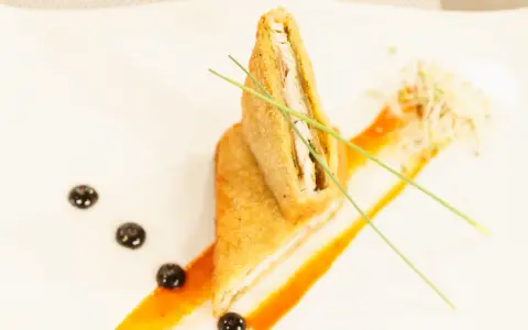Un crujiente san jacobo, cortado por la mitad, reposa sobre una salsa naranja. Brotes y puntos oscuros decoran el plato.