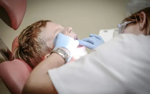Chłopiec siedzi na fotelu dentystycznym podczas wizyty u dentysty. Dentysta w rękawiczkach bada jego zęby.