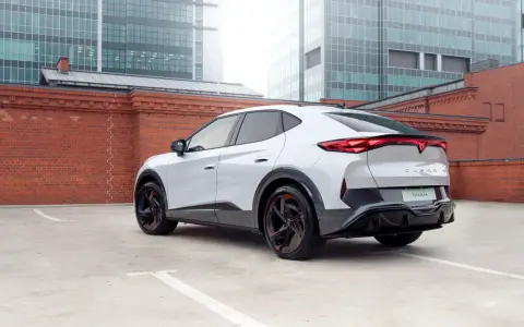 Biały elektryczny SUV Cupra Tavascan z czerwonymi akcentami na felgach, zaparkowany na tle ceglanego muru i nowoczesnych budynków.