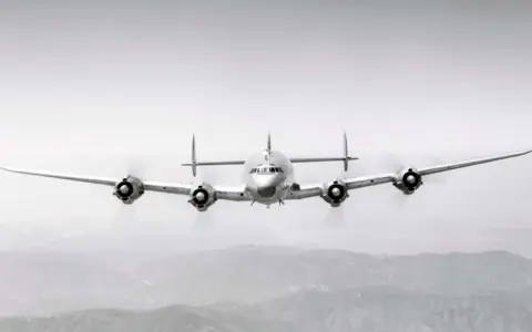 Ein vier-motoriger Propellerflieger mit markantem Leitwerk fliegt über bergiger Landschaft.