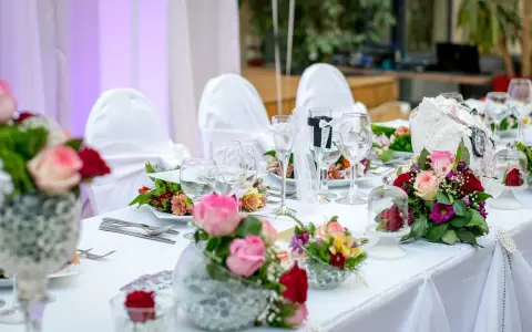 Elegancki, samodzielny wystrój sali weselnej: białe pokrowce na krzesła, bukiety róż i goździków, kieliszki do wina i szampana.
