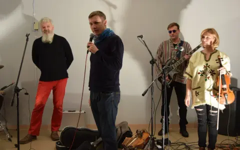 Orkiestra Świętego Mikołaja przygotowuje się do występu. Wokalista z mikrofonem, gitarzysta, skrzypaczka i brodaty mężczyzna w czerwonych spodniach.