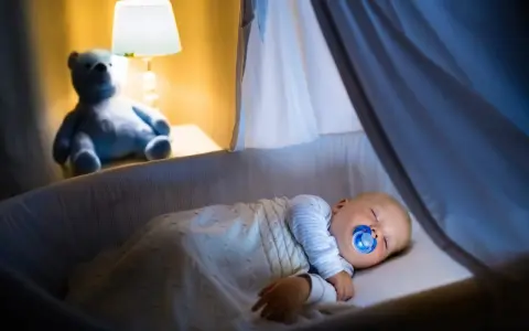 Bambino dorme sereno nella culla, con ciuccio, sotto una coperta calda. Un orsacchiotto veglia, simbolo di addormentamento autonomo.