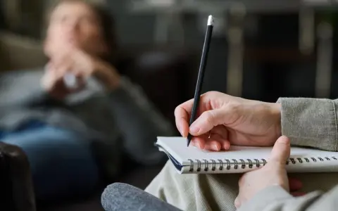 Psycholog pomaga zrozumieć siebie i swoje problemy. Na zdjęciu widać rękę terapeuty piszącą w notatniku, podczas gdy pacjent leży na kanapie.