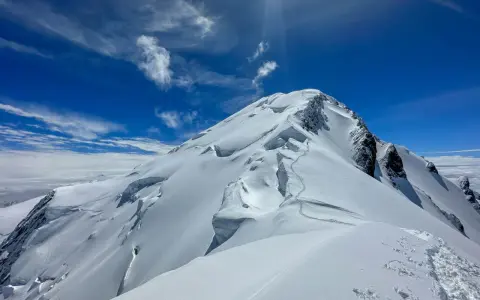 Śnieżna trasa na Mont Blanc, z widocznymi szczelinami lodowcowymi i śladami wspinaczy. Błękitne niebo z chmurami.