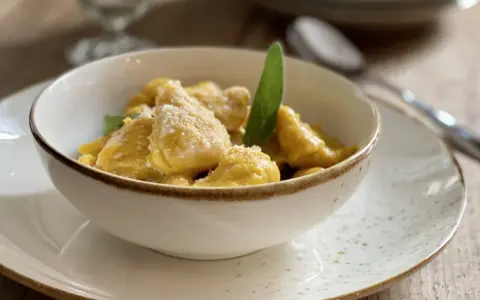 Gnocchi di zucca dorati e soffici, conditi con parmigiano e una foglia di salvia, serviti in una ciotola bianca su un piatto maculato.