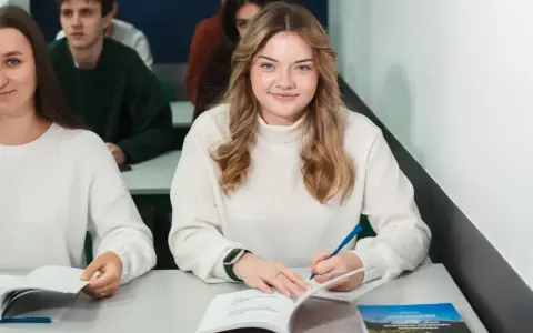 Uśmiechnięta studentka pisze notatki, zastanawiając się, ile wynosi stypendium na studiach. W tle inni studenci.