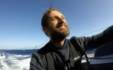 Un homme de mer souriant, le visage tourné vers le ciel bleu, navigue sur un voilier. L'océan agité et le soleil radieux témoignent de son aventure.