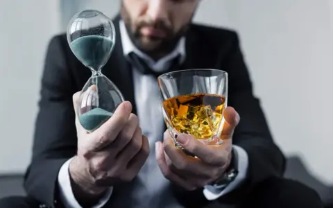 Mann im Anzug hält Sanduhr und Whiskyglas. Die Frage, ob kann Whisky schlecht werden, wird durch die vergehende Zeit und den edlen Tropfen symbolisiert.