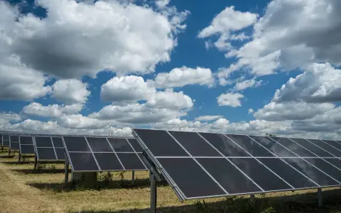Rzędy paneli fotowoltaicznych 50kW na tle błękitnego nieba z białymi chmurami. W oddali widać turbiny wiatrowe.