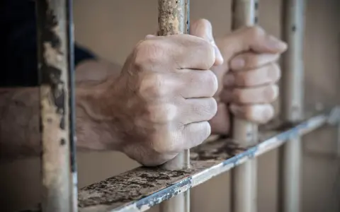 Dłonie zaciskają się na kratach aresztu śledczego. Widok zza prętów.