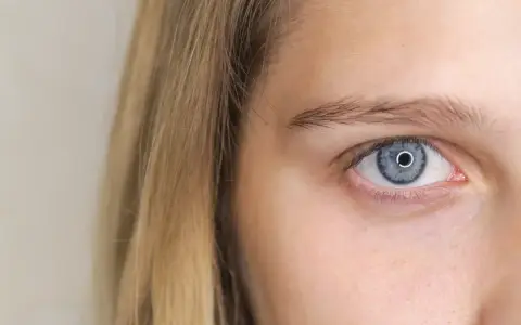 Błękitne oko z jasną tęczówką, gdzie szpilkowate źrenice charakterystyczne są. Blond włosy opadają na twarz.