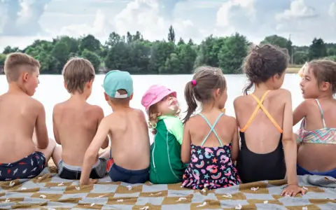 Dzieci siedzą na kocu nad jeziorem, ciesząc się fajnymi wczasami. Jedno z nich ma lody na twarzy.