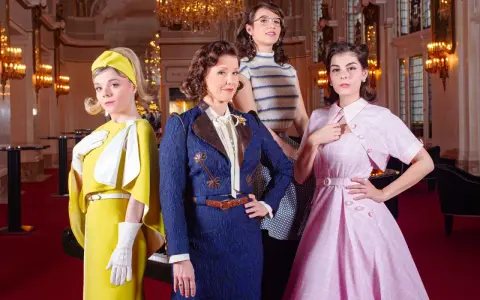 Vier Frauen in stilvollen Outfits, die an die Kleiderordnung des Theater des Westens erinnern, posieren vor einem opulenten Hintergrund.