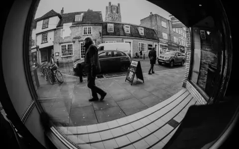Calle con edificios, coches y gente. Un hombre camina frente a un cartel de "Caffè Nero" con efecto ojo de pez.