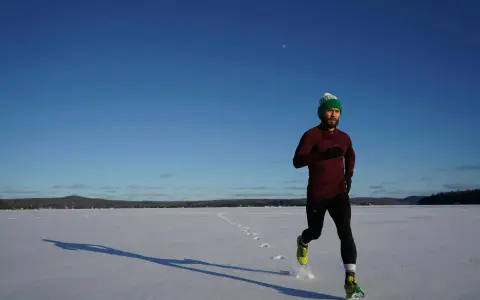 Mann mit grüner Mütze läuft im Winter über eine verschneite Fläche, die Sonne wirft lange Schatten.