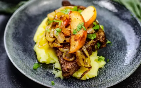 Gedünstete Leber mit Apfelspalten, Zwiebeln und Kartoffelpüree auf einem dunklen Teller.