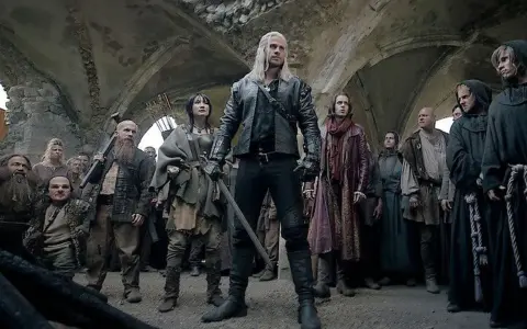 Geralt z Rivii stoi pośród tłumu, gdy ścieżki przeznaczenia w Wiedźminie splatają się. Widać krasnoludy, ludzi i zakapturzonych mnichów.