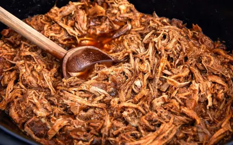 En stor gryta med saftig, långkokt pulled pork i en mörk sås. En träsked vilar i den läckra pulled pork grytan.