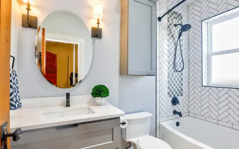 Une petite salle de bain moderne avec baignoire, lavabo gris, miroir rond et carrelage chevrons. Une belle idée pour optimiser l'espace.