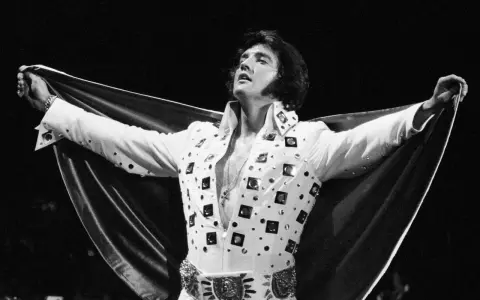 Elvis Presley, der unzählige Tonträger verkaufte, posiert in einem glitzernden weißen Anzug mit ausgebreitetem Umhang.